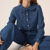 Modèle assise portant la blouse en denim fluide Lenora DES PETITS HAUTS avec col bandana à nouer avec un jean et des chaussures metallisées