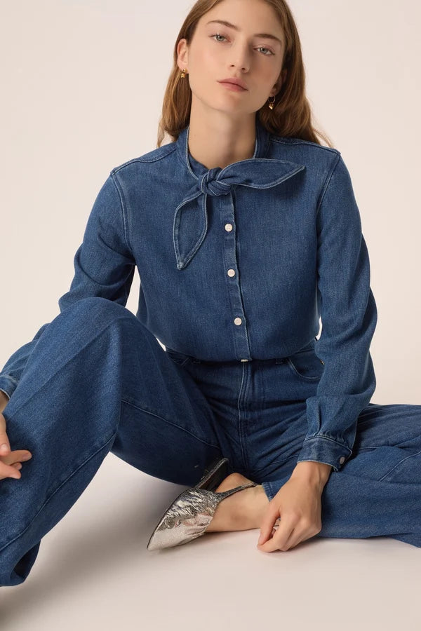 Modèle assise portant la blouse en denim fluide Lenora DES PETITS HAUTS avec col bandana à nouer avec un jean et des chaussures metallisées