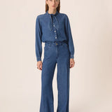 Modèle portant la blouse en jean bleu Lenora DES PETITS HAUTS avec le col noué sur le côté et un jean avec des chaussures métallisés