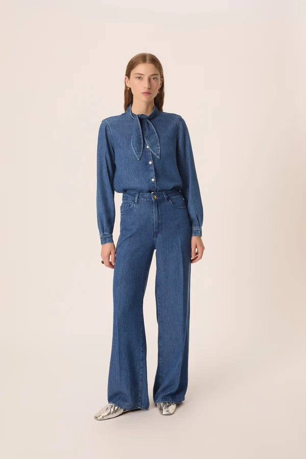 Modèle portant la blouse en jean bleu Lenora DES PETITS HAUTS avec le col noué sur le côté et un jean avec des chaussures métallisés