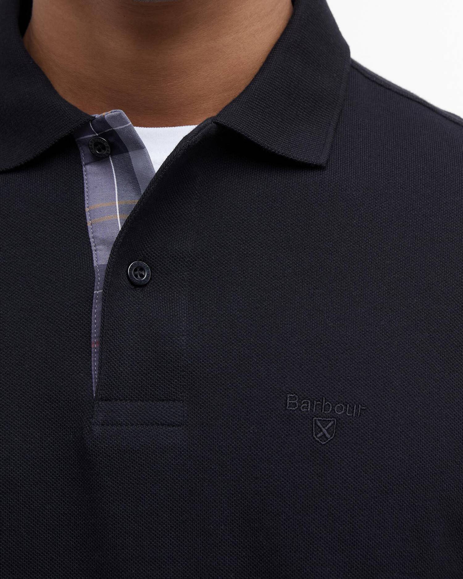 Gros plan sur le col du polo Essentiel noir Barbour, la doublure tartan et le logo brodé sur la poitrine